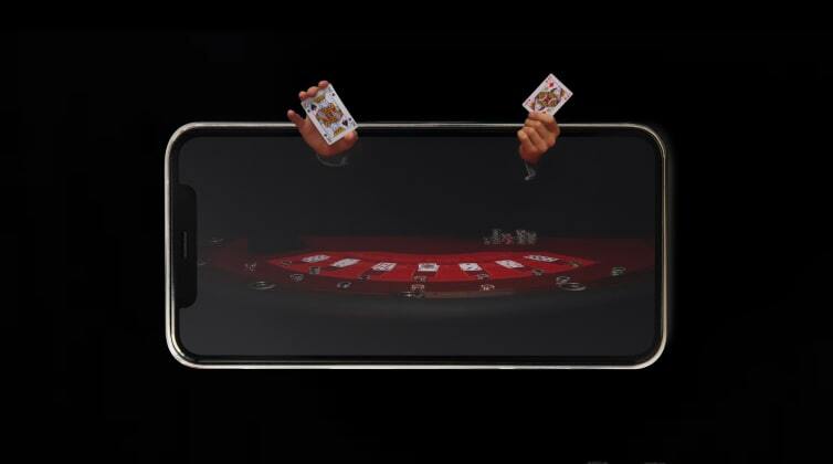 Live casino poker game on a mobile phone screen, with hands holding cards appearing to come out of the device, creating an immersive gaming experience.